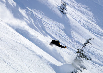 A skier and snowboarder conquering the pristine, snowy slopes at Eaglecrest in Juneau, Alaska. A snowmobile waits nearby, while a chalet stands in the background amidst the winter sports scene.