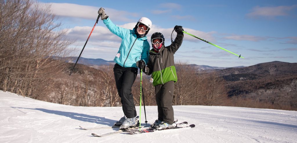 Winter scene at Middlebury Snowbowl, Vermont featuring a skier descending a slope, with a ski lift visible in the background. Other skiers, possibly a family, are seen further down the slope.