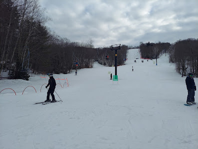 A skier descends a snow-covered slope at Middlebury Snowbowl in Vermont with a ski lift and a chalet visible in the background. The scene encapsulates a typical day in a winter sports resort.