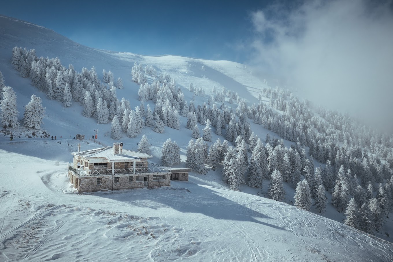 Spectacular view of Ski Center in Chelmos, Kalavrita, displaying a buzzing winter sports scene. A mountain hut rests amidst a mesmerizing winter landscape with snow-clad slopes under a clear blue sky.