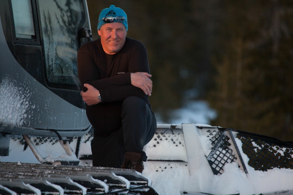 A skier glides through the snowy landscape of Sjusjøen in Lillehammer, Norway. A snowmobile and snowboarder are also seen actively embracing winter sports. In the distance, a chalet nestles amid the winter wonderland.