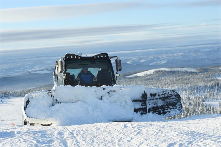 A winter sports scene at Sjusjøen in Lillehammer Norway. A snowmobile and a skier are present against the backdrop of a chalet and a ski lift blanketed in snow.