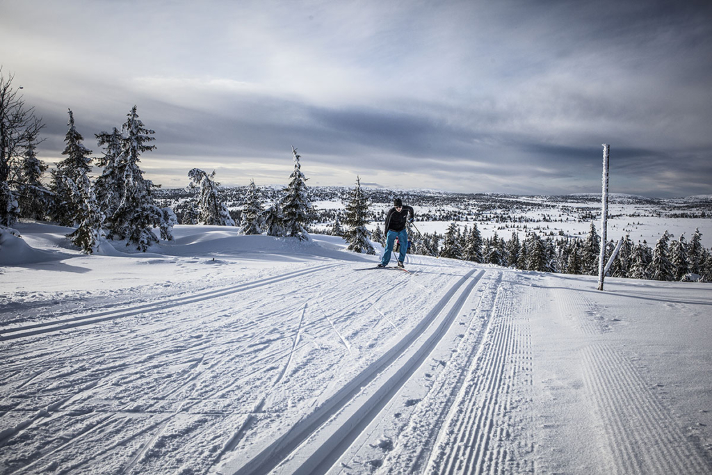 Winter sports scene at Sjusjøen in Lillehammer, Norway, featuring a lone skier gliding across a snow-covered landscape with a charming challet and the rest of the ski resort in the background.