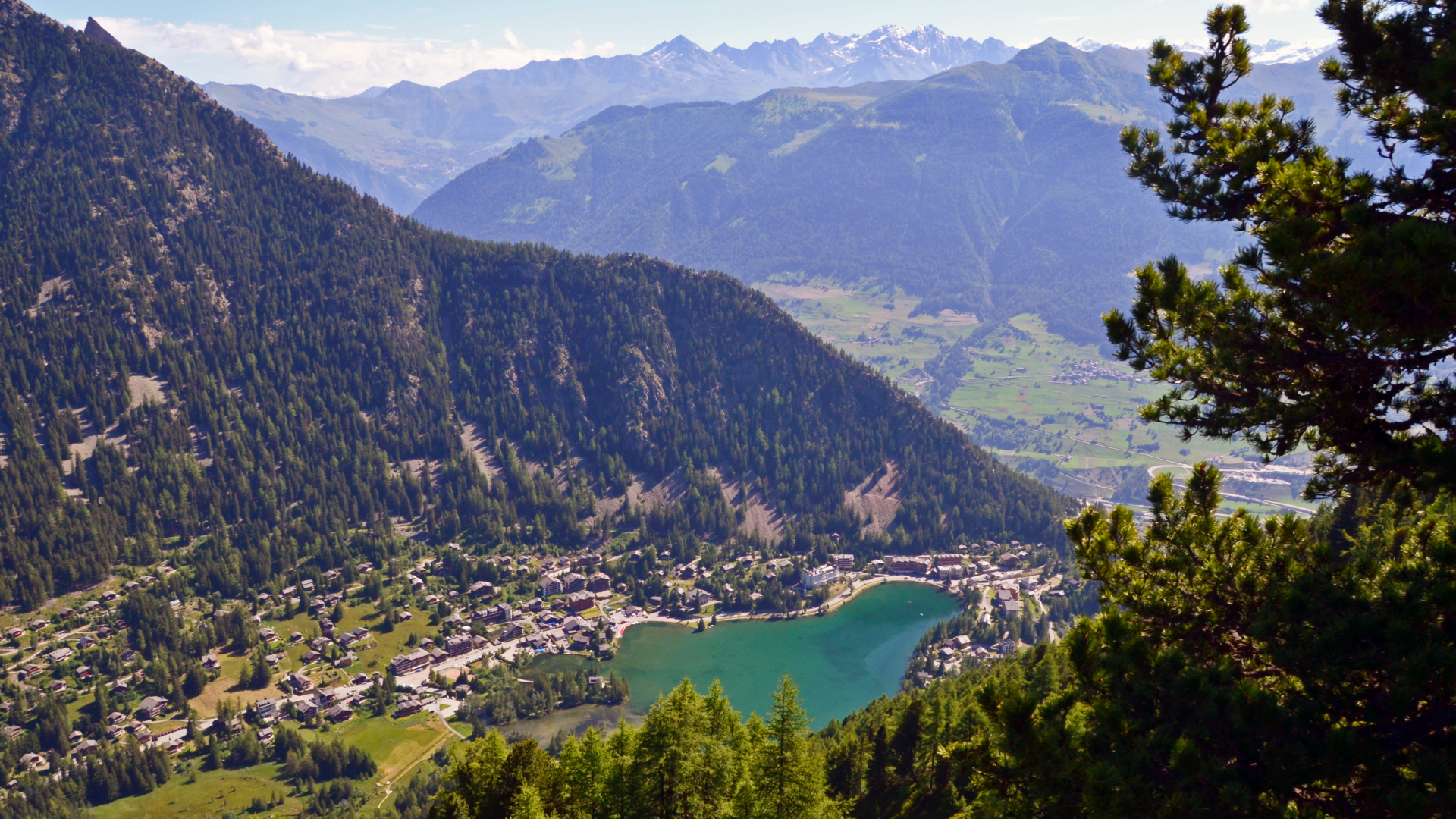 Charming chalet nestled near a tranquil lake in the scenic town of Champex-Lac, Switzerland. Stunning mountains provide a grand backdrop under the clear, sunny skies. Hint of a ski resort suggests active outdoor enjoyment.