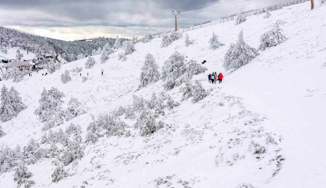 Winter sports scene at Puerto Navacerrada in Madrid, Spain. Image features a bustling ski resort with a ski lift running up the snow-covered slopes, amidst stunning winter scenery, under a clear sky.