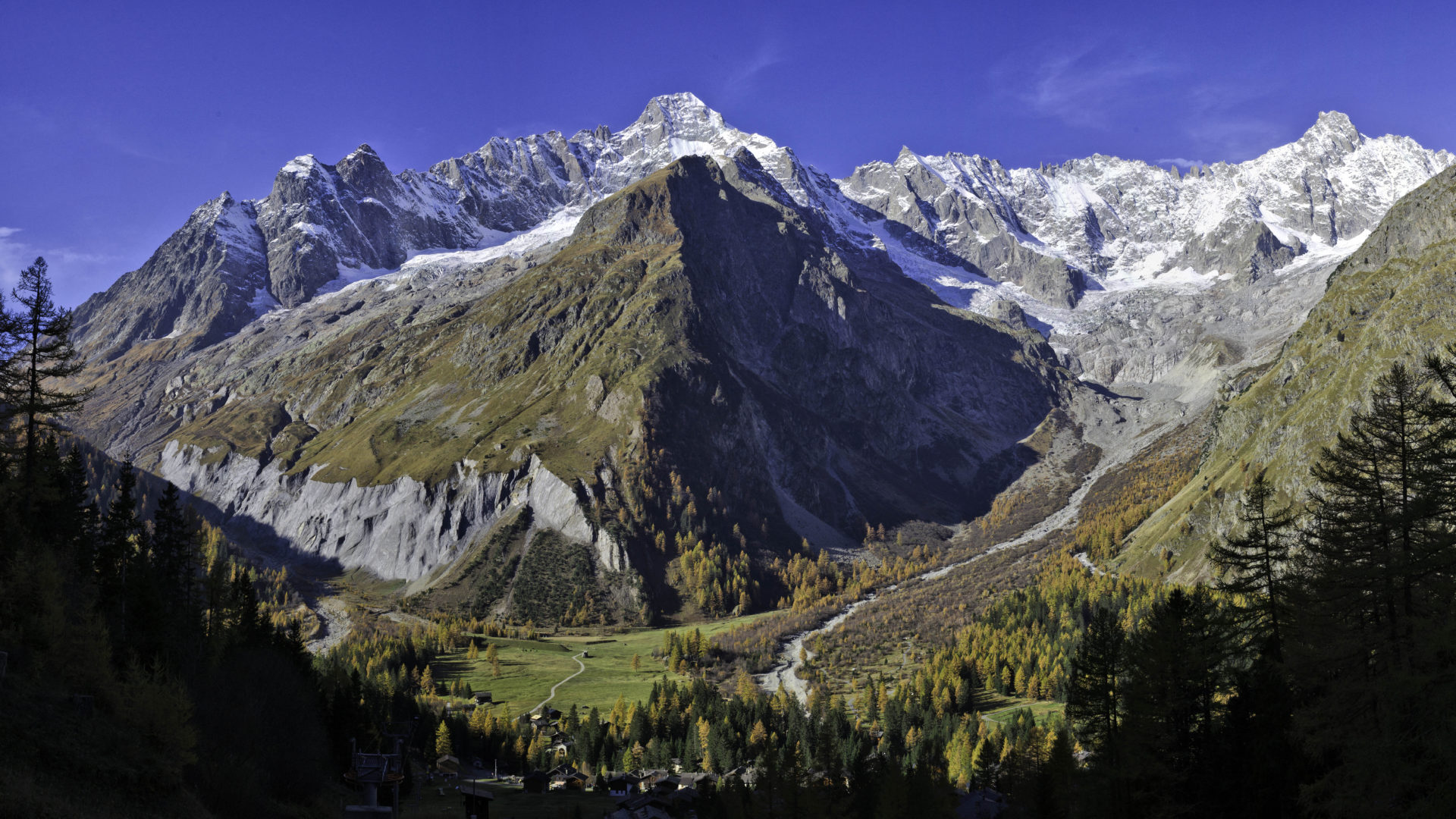 A picturesque Swiss chalet in La Fouly, Valais, featuring a breathtaking view of a towering mountain. It exudes warmth and coziness, nestled at the foot of the snow-covered slope, hinting at a nearby ski resort.