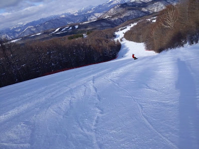 A skier is gliding down the snowy slopes of Hunter Mountain Shiobara in Japan, with a ski lift and chalet in the distant backdrop contributing to a lively winter sports scene.