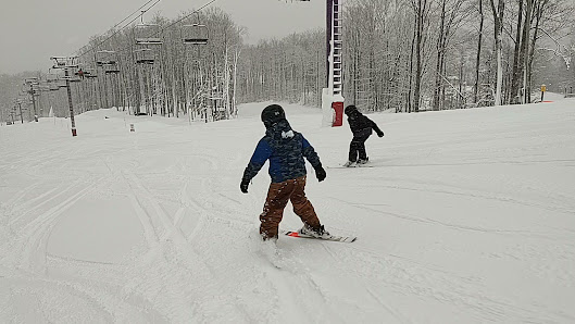 A snowboarder skillfully descending Schuss Mountain at Shanty Creek Resorts in Mancelona, Michigan. The mountain is blanketed in a thick layer of snow under a clear, blue sky.