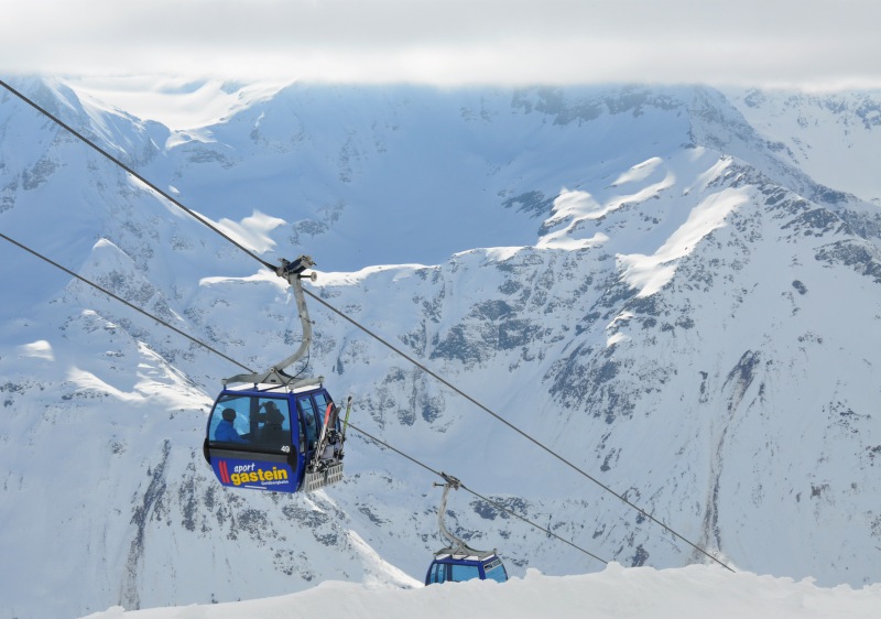 A picturesque Austrian ski resort sight captured at Bad Gastein showing bustling ski activity. The scenery includes a ski lift transporting adventure-seekers a skier gliding down and a charming chalet in the snowy backdrop.