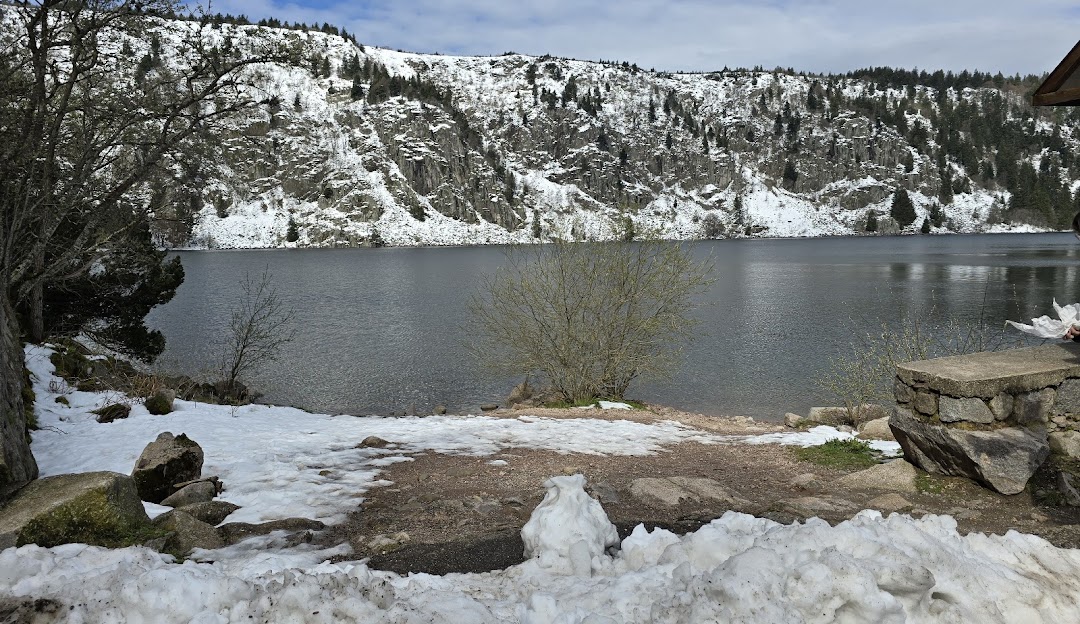 Photo of a charming chalet overlooking the serene Lac Blanc in Haut-Rhin, France. A picturesque winter scene unfolds with snow-covered mountains in the background.