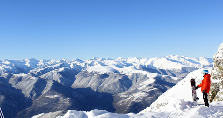 A chalet at Les Monts d'Olmes ski resort in Ariège, France, set against a scenic mountain backdrop. A skier is visible in the winter sports scene.