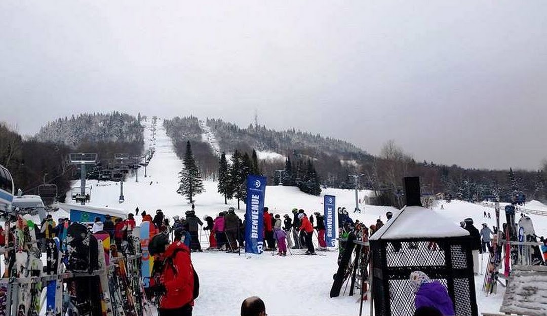 Skiers and snowboarders enjoy the snowy slopes at Ski La Réserve in Quebec, Canada. The scenic landscape is dominated by a ski resort, various winter sports in progress, and a ski lift transporting visitors up the mountain.