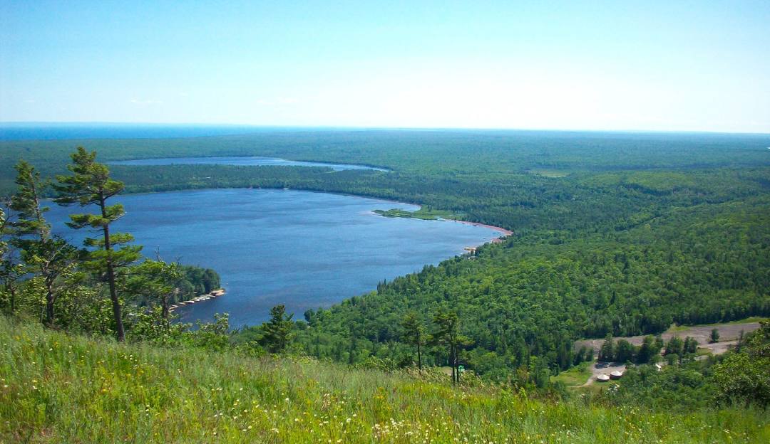 View of a serene lake nestled beneath Mt Bohemia in Mohawk, Michigan. A challet can be seen off to the side, with a beautiful clear sky reigning above the scene on a sunny day.