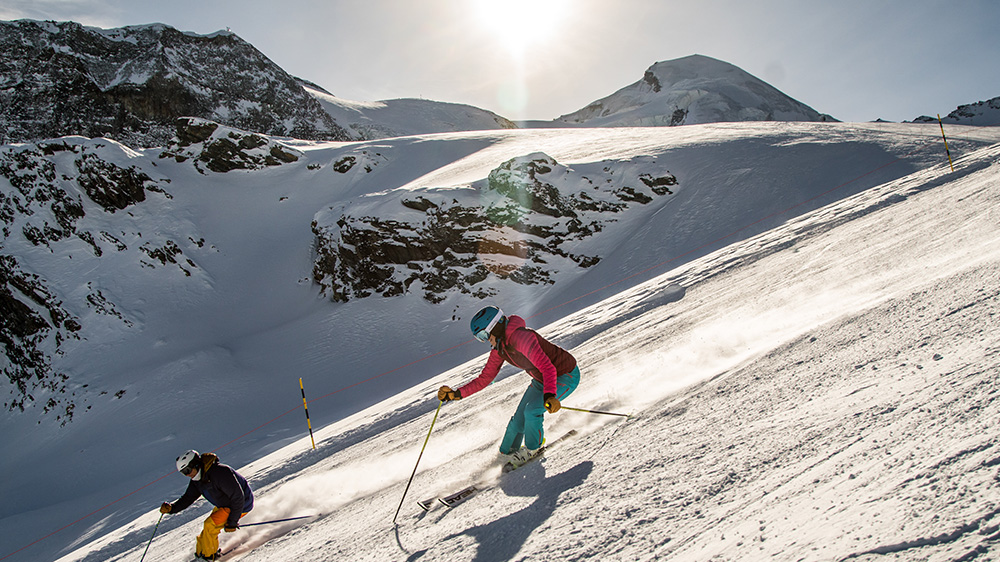 A skier is carving down the snowy slopes of Saas-Almagell in Switzerland with a group of people possibly a family skiing further behind. A charming chalet is nestled in the background contributing to this idyllic winter sports scene.