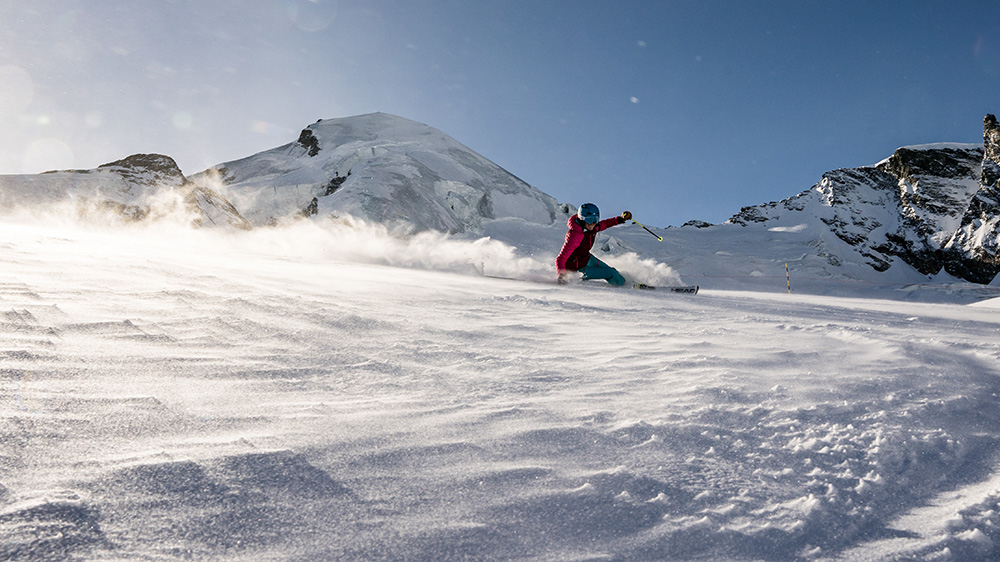 A skier in action at the scenic Heidbodme – Saas-Almagell in Switzerland, with a snowboarder and winter sports enthusiasts in the backdrop. A charming chalet and ski resort complete the snowy landscape.