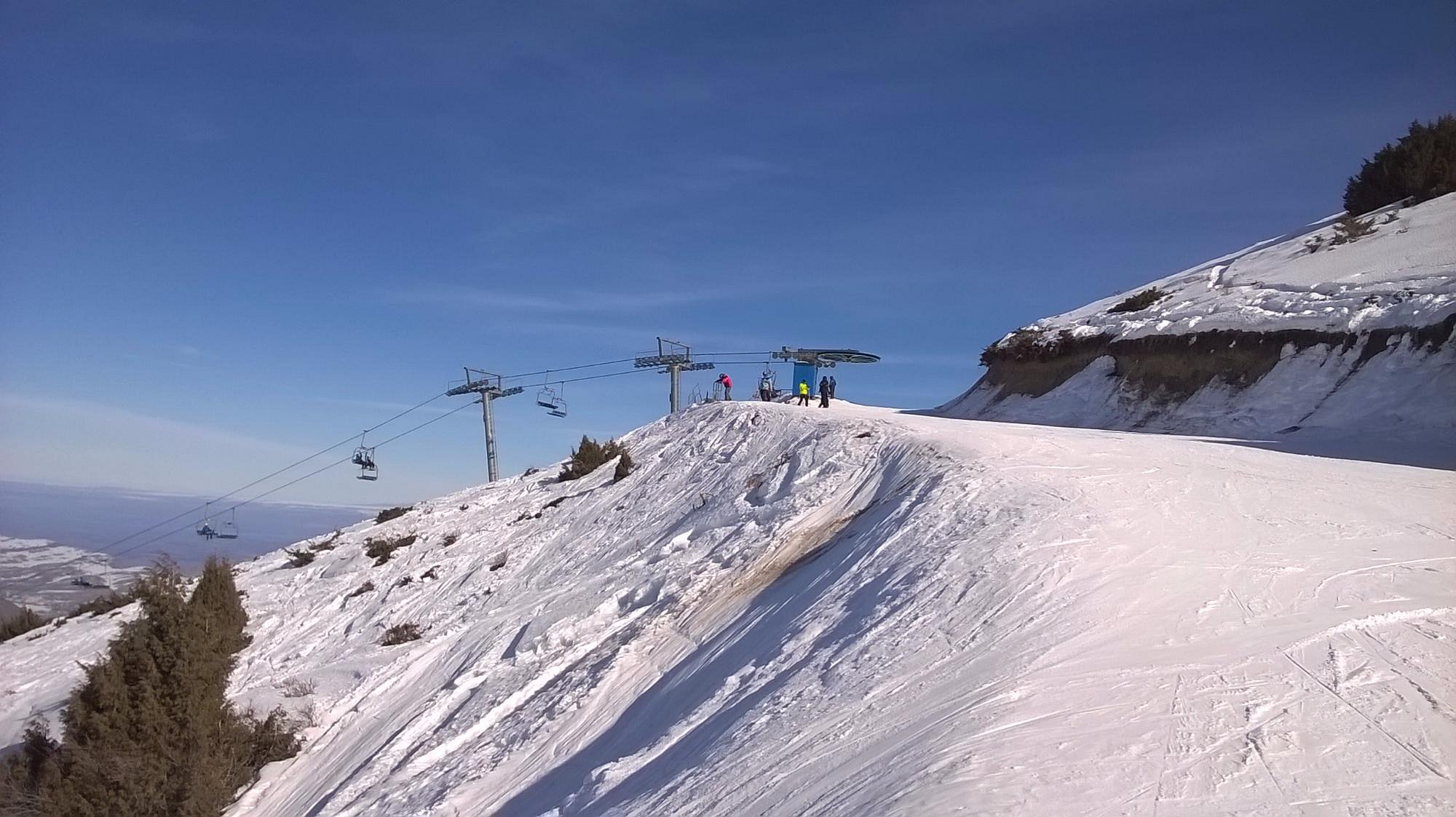 Ski scene at Chunkurchak, Kyrgyzstan, depicting a bustling ski resort with skiers dotting the slopes. A ski lift can be spotted, hinting towards a ski chalet partially hidden in the background.