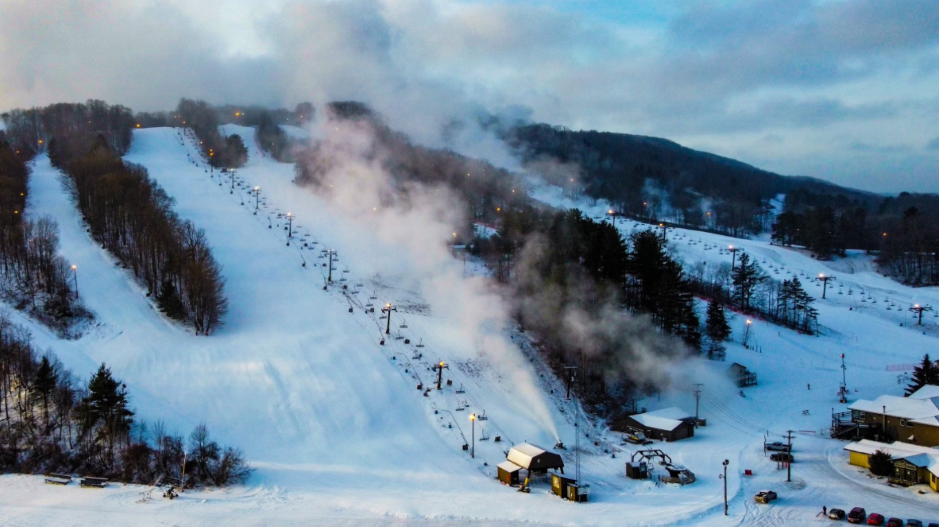 Winter scene at Swain ski resort in New York, USA, featuring a bustling ski slope with skiers, a ski lift moving in the background and serene, snow-covered scenery.