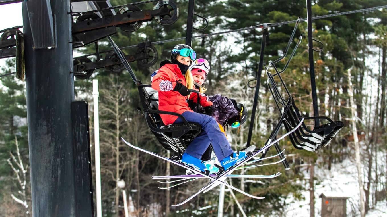 A winter sports scene at Swain ski resort in New York, featuring a skier and a snowboarder on a snowy slope and a ski lift in the background.