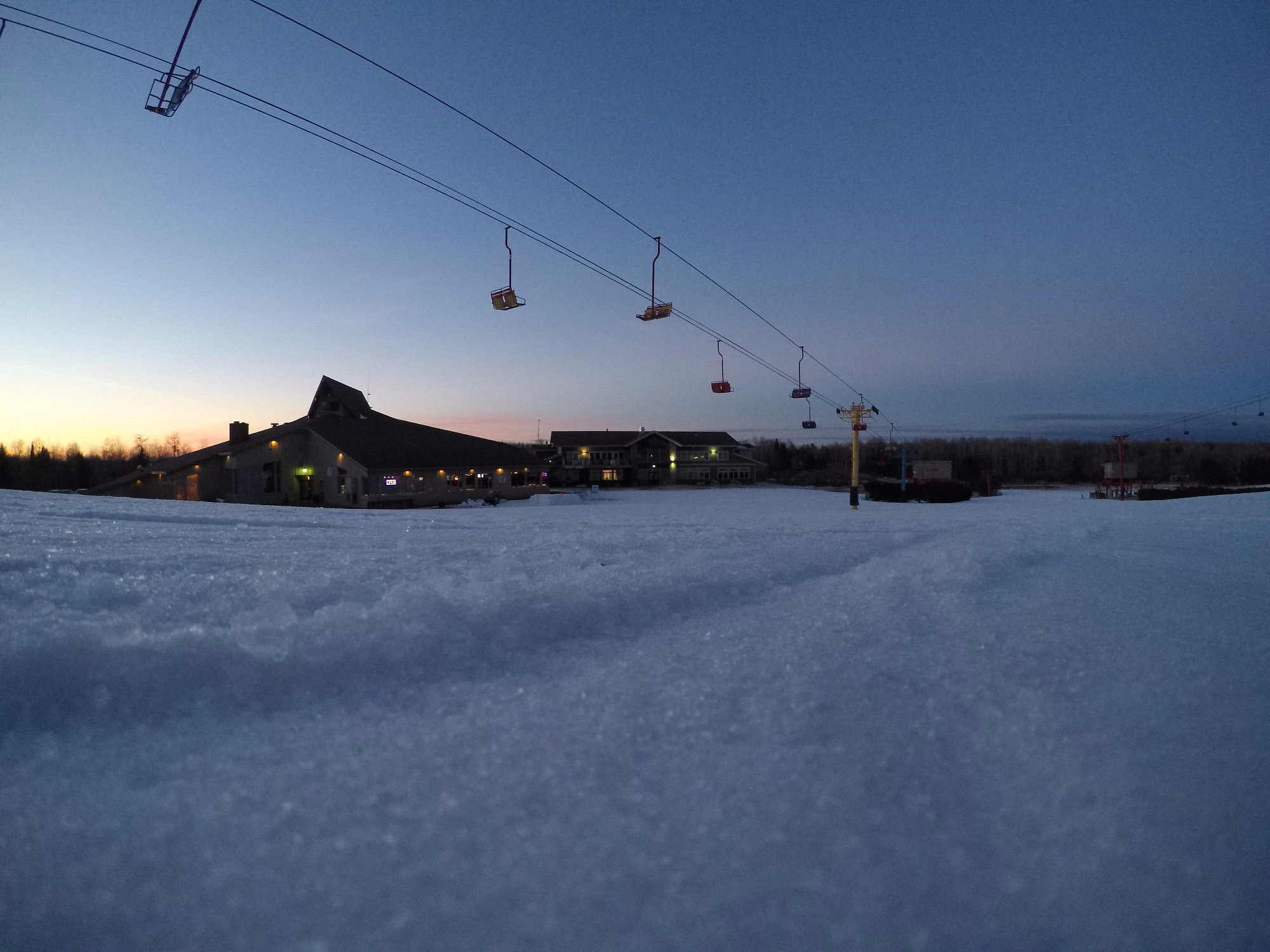 A view of Big Powderhorn Mountain in Michigan features a ski lift ascending the snowy slope, dotted with winter sports enthusiasts. Surrounding structures hint at a ski resort and chalet.