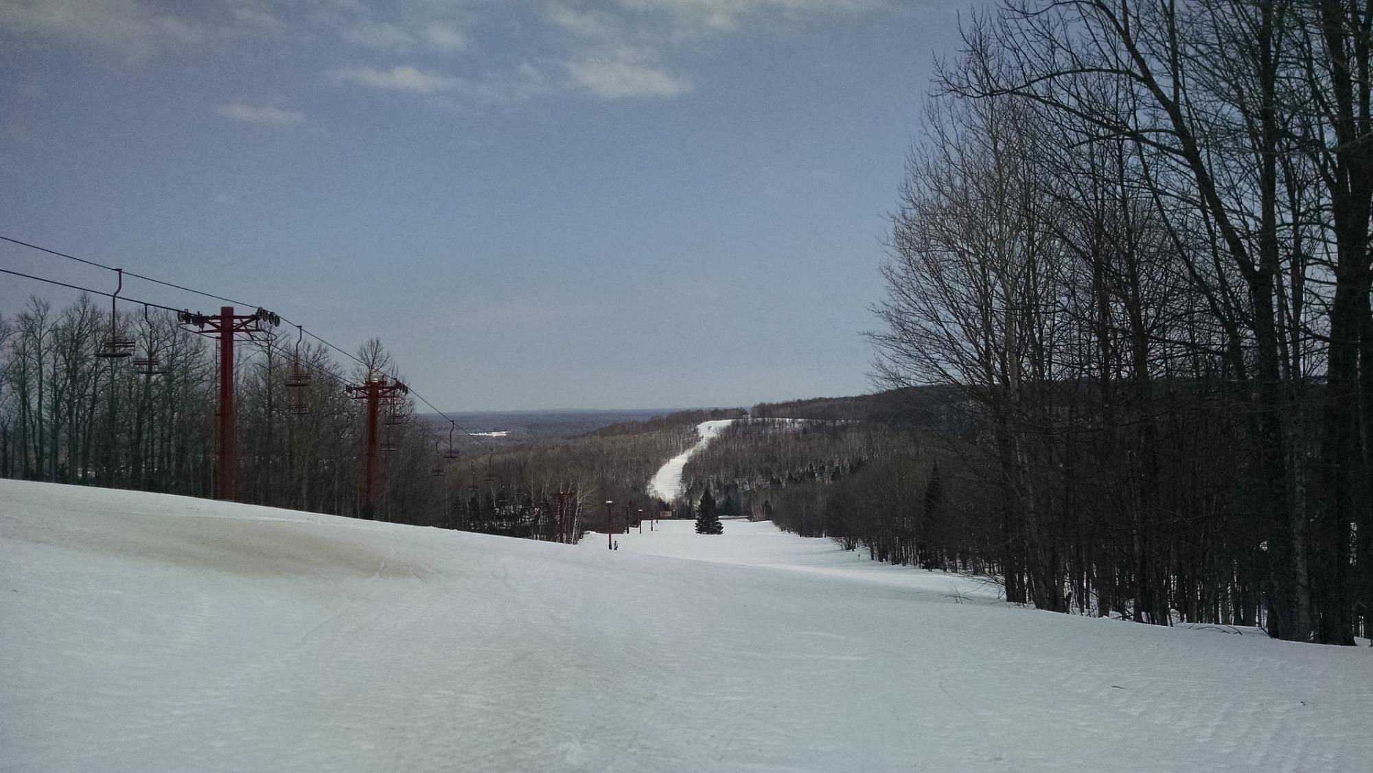 Snow-covered ski slopes at Big Powderhorn Mountain in Bessemer Michigan bustling with skiers. A ski lift carries more enthusiasts up the mountain while a cozy chalet peeks from the corner.