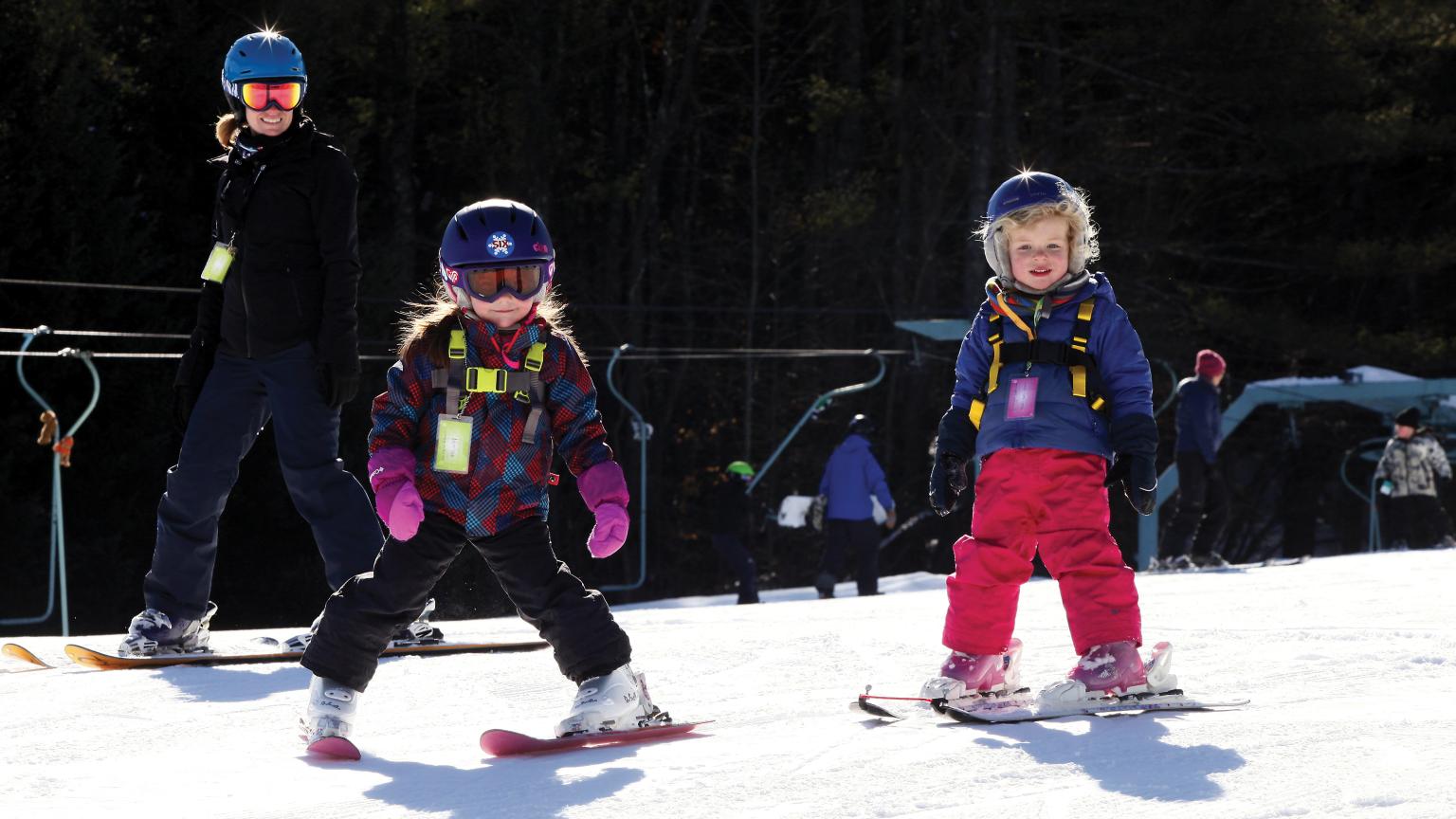 Winter scene at Saskadena Six, Vermont, featuring a family enjoying skiing together with a child learning. A ski lift is visible in the background.