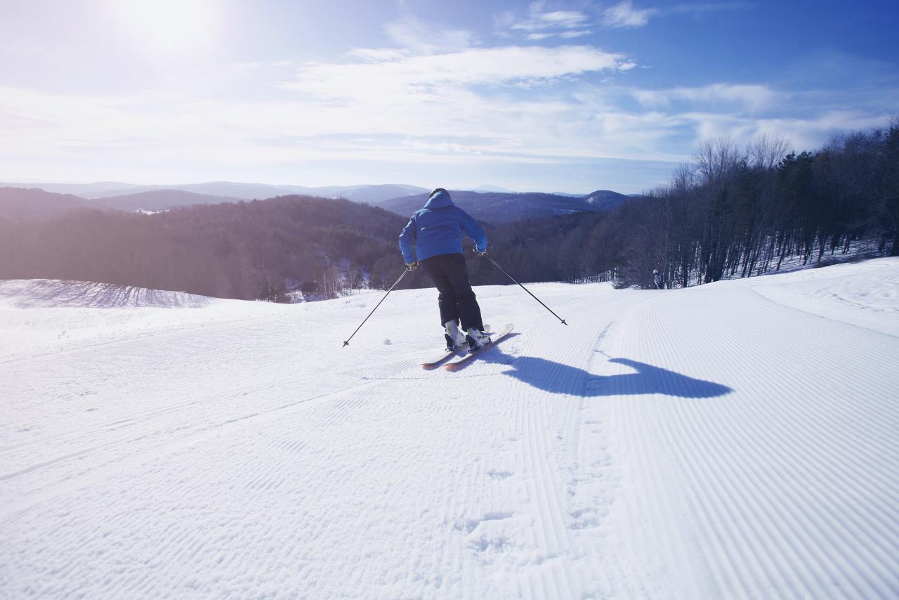 A skier glides down the snowy slopes at Saskadena Six in South Pomfret, Vermont. Nearby, a family enjoys a group ski, with a cozy chalet nestled in the background, providing warmth and comfort in contrast to the snowy expanse.