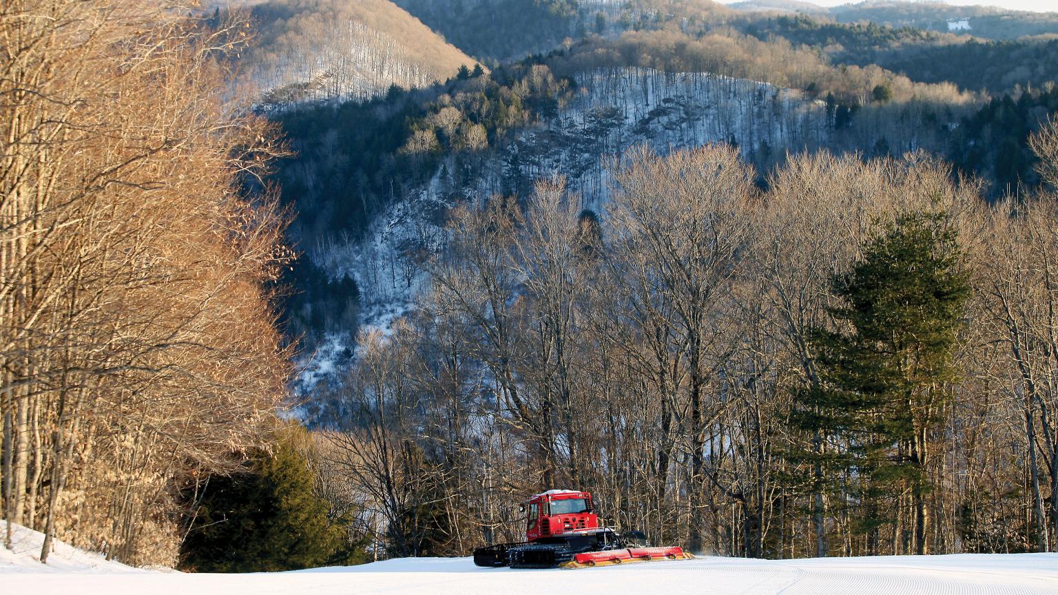 A winter sports scene at Saskadena Six in Vermont featuring a ski lift transporting visitors towards a warm welcoming chalet at a ski resort. Fresh snow can be seen with a parked snowmobile nearby.