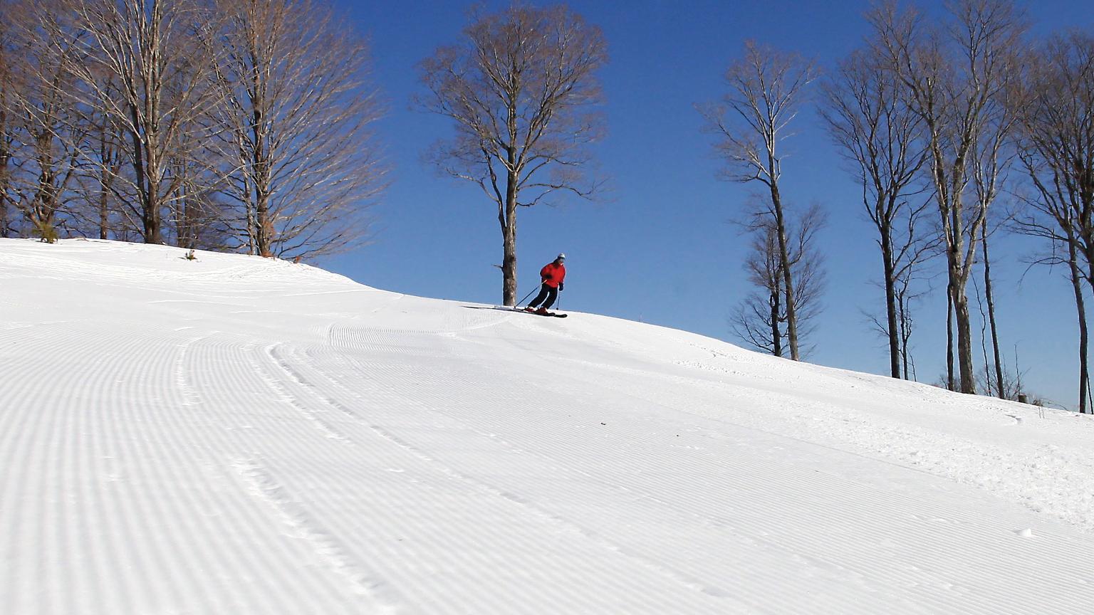 A skier is enjoying a brisk day at Saskadena Six in South Pomfret, Vermont. The winter sports scene is set at a ski resort, where a faint image of a chalet can be seen in the background.