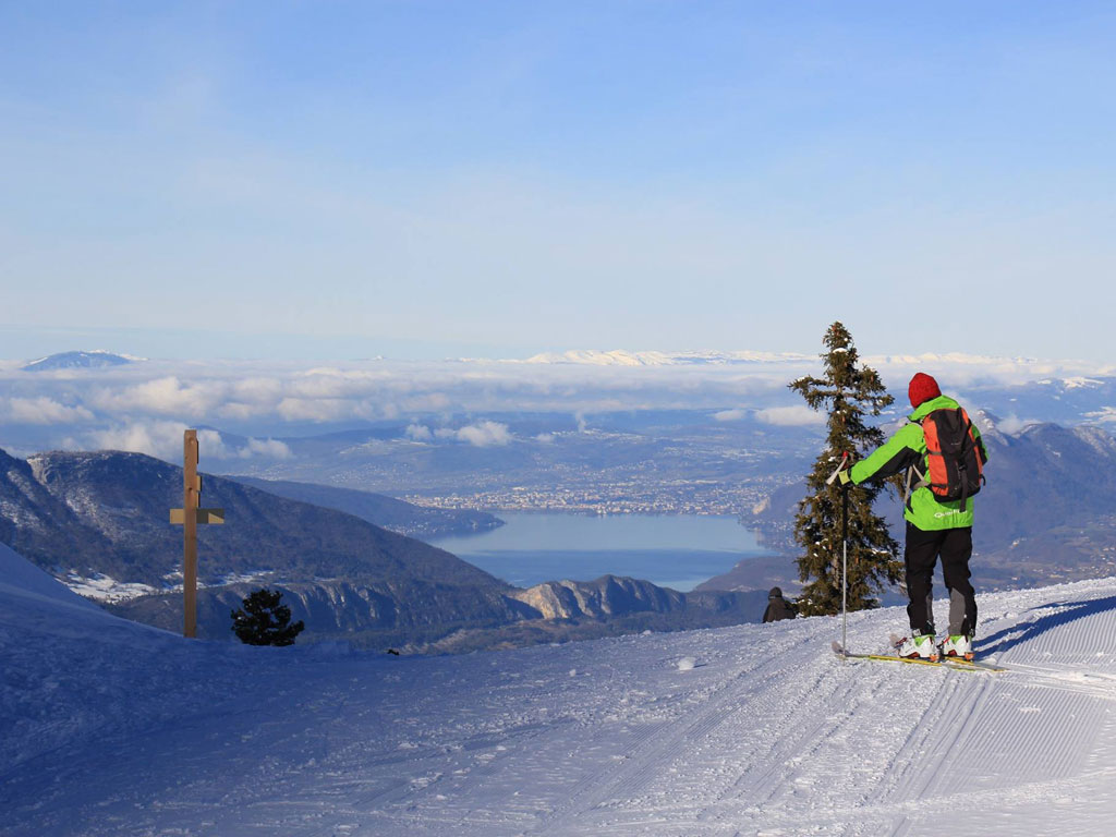 Winter sports scene in La Sambuy-Seythenex, France, featuring a skier and a snowboarder descending the snowy slopes, with a ski lift present in the background.