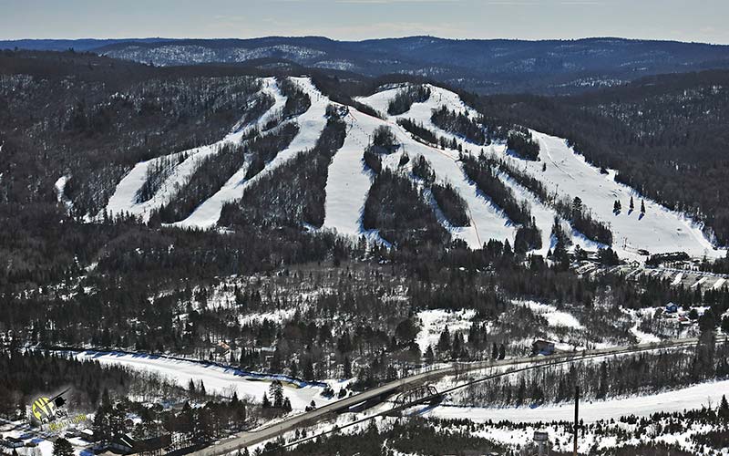 View of Searchmont ski resort in Ontario, featuring snow-covered slopes bustling with winter sports activity. Visible elements include a ski lift and a skier enjoying the pristine conditions.