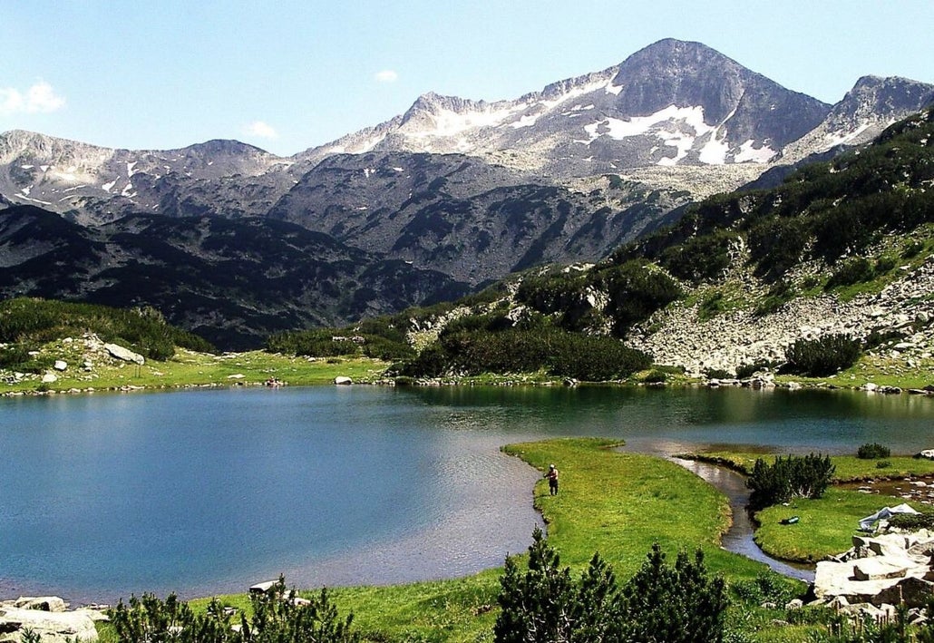 Winter scene in Bansko, Bulgaria featuring a calm lake, a cozy challet by it, surrounded by vast snowy mountains, home to a welcoming ski resort.