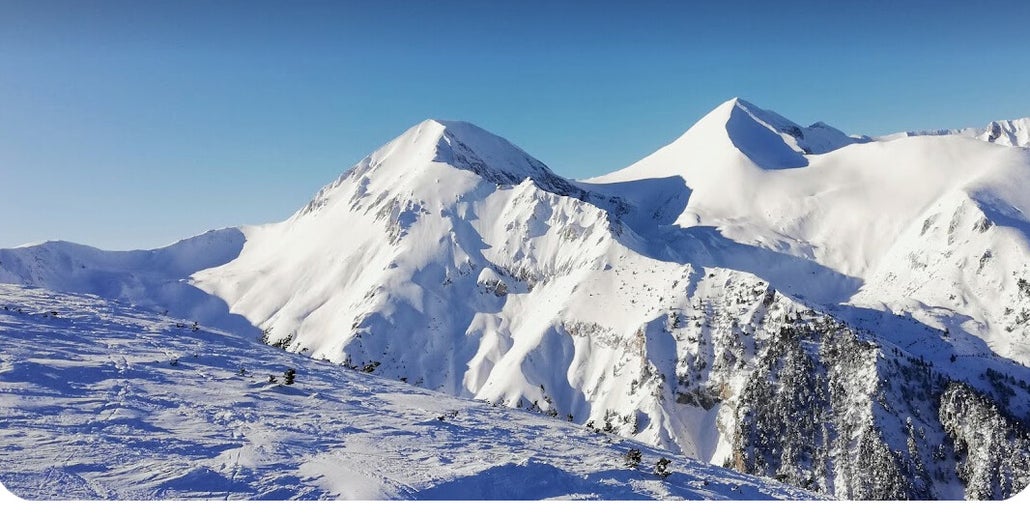 View of Bansko, Bulgaria featuring a prominent, snow-covered mountain, with a ski resort and individuals participating in winter sports sprinkled across the scenic winter landscape.