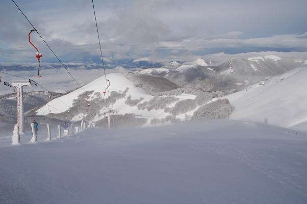 A picturesque scene of Monte Terminillo's ski resort in Italy featuring a charming chalet and ski lift. Skiers enjoy the snow-covered slopes under a bright sky.