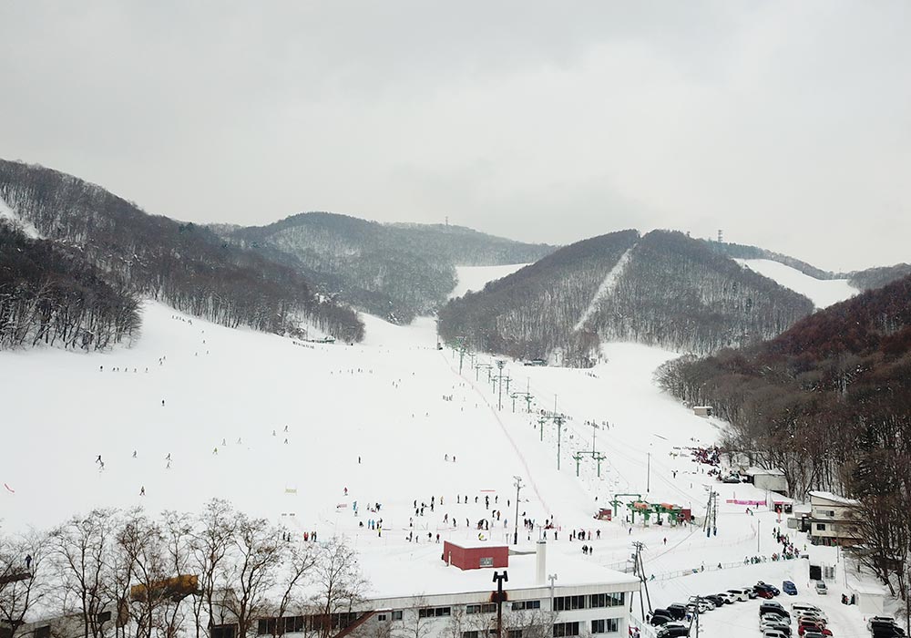 A spectacular winter view at Moiwayama Ski Jominamishamen in Japan, featuring a bustling ski resort in the backdrop of snow-covered slopes. The scene is alive with winter sports activity.