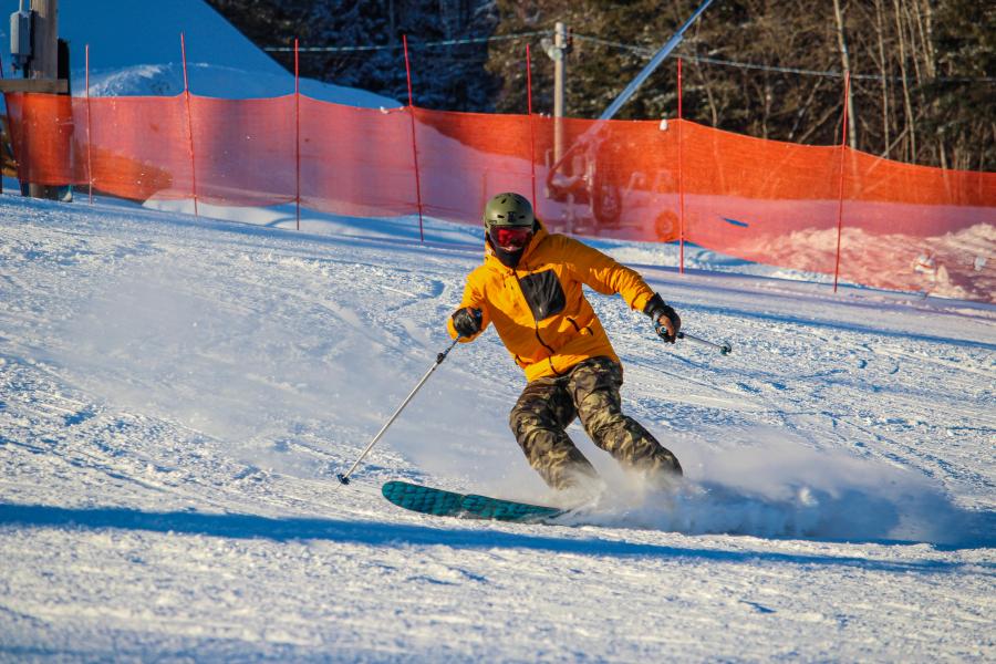 A winter sports scene at Mont Farlagne in Saint-Jacques, New Brunswick, Canada, featuring a skier and a snowboarder on the snowy slopes with a chalet and the winter sports center in the background.