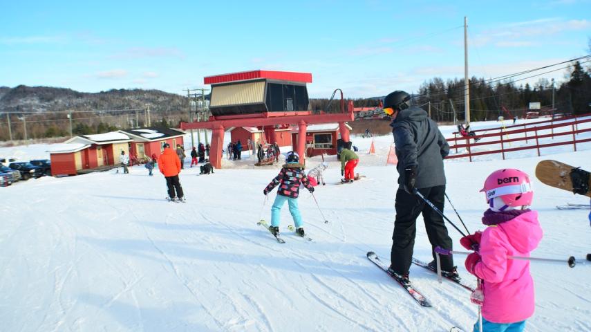 Winter sports scene at Mont Farlagne in Canada, featuring a bustling sports centre, a ski challet and several skiers, including a family enjoying their ski down the snowy mountain.