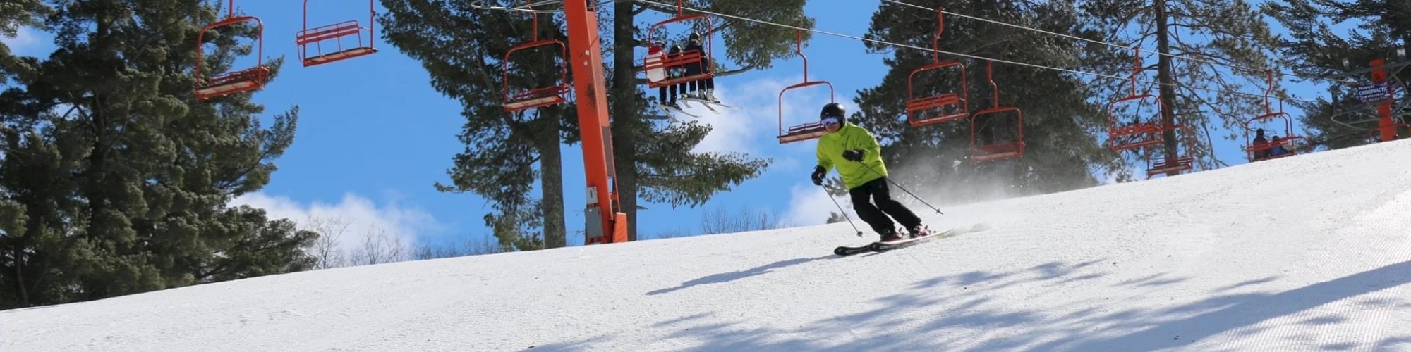 A skier gliding down the slopes of Pine Mountain in Iron Mountain Michigan. Nearby a ski lift is visible taking enthusiastic winter sports enthusiasts up to the peak. A charming chalet peeks out from the snowy landscape.