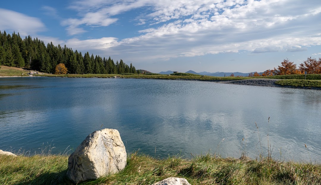 A serene view of a tranquil lake in Annaberg, Austria, nestled among breathtaking mountain scenery, with a charming chalet in the distance, all under a bright sunny sky.