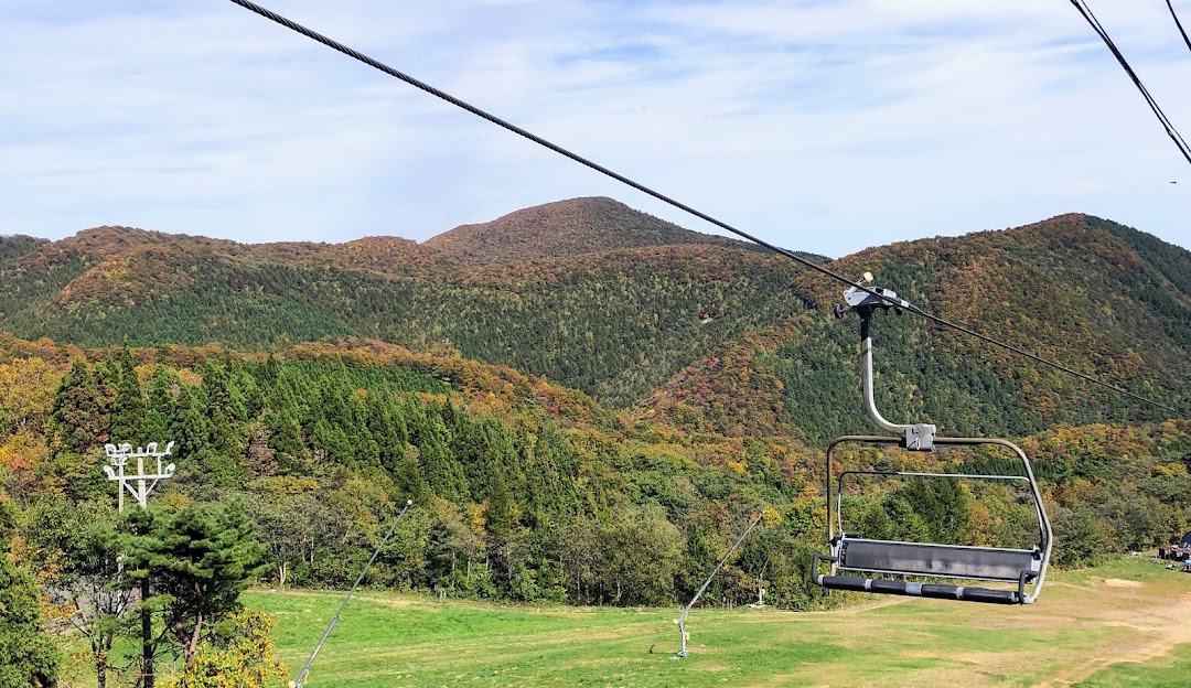 A ski lift ascends up a mountain in Spring Valley Izumi Kogen Sendai a ski resort in Japan. A quaint chalet settles in the backdrop under a serene sky with a lone skier navigating the terrain.