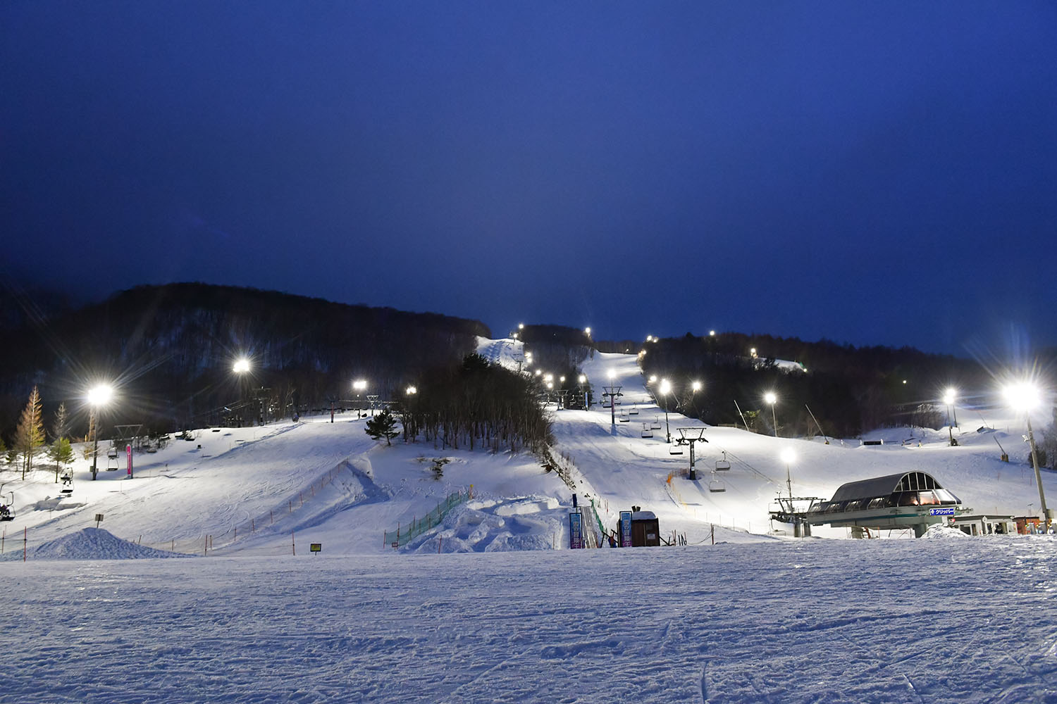 View of Spring Valley Izumi Kogen Sendai, a bustling ski resort in Japan. The scene depicts snow-covered slopes, skiers in action, and a ski lift operating in the background.