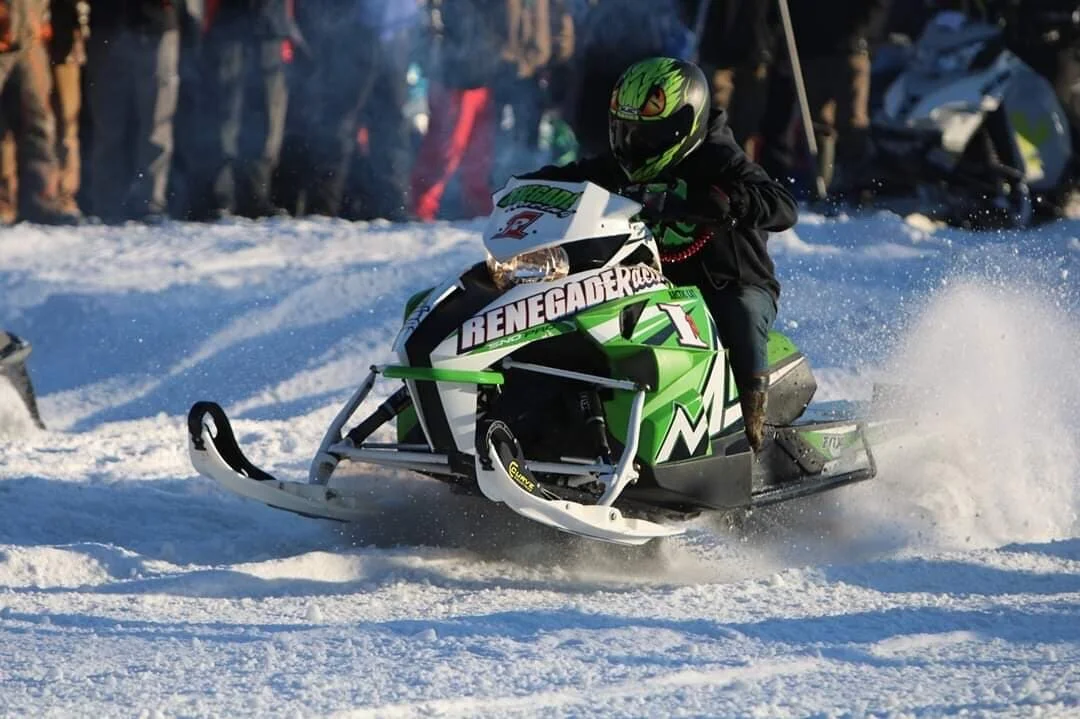 A snowmobiler enjoying a ride on the snowy trails of Royal Mountain in Johnstown New York. The surroundings are a winter sports haven with distant glimpses of a skier and a chalet.
