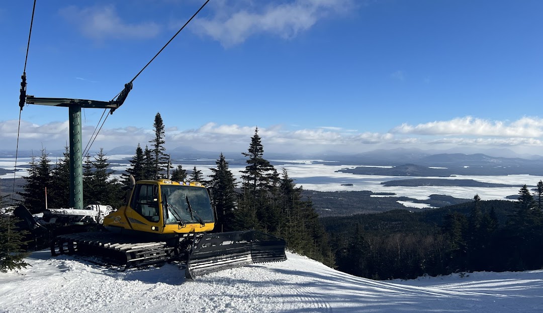 View from Big Moose Mountain in Maine, USA, featuring a ski lift ascending the mountain. The scene captures the essence of winter sports at a ski resort, with a skier and snowmobile in the distance.
