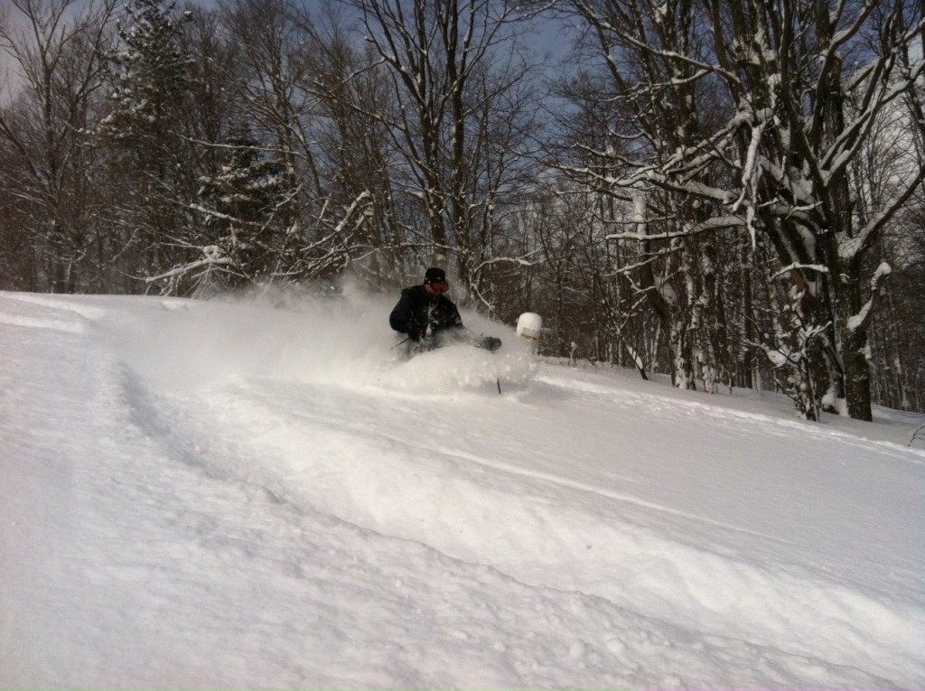 A skier gliding down the snowy slopes at Snow Ridge, The Adirondacks, Turin, NY, with a snowmobile and a chalet spotted in the background, contributing to the exhilarating winter sports scene.
