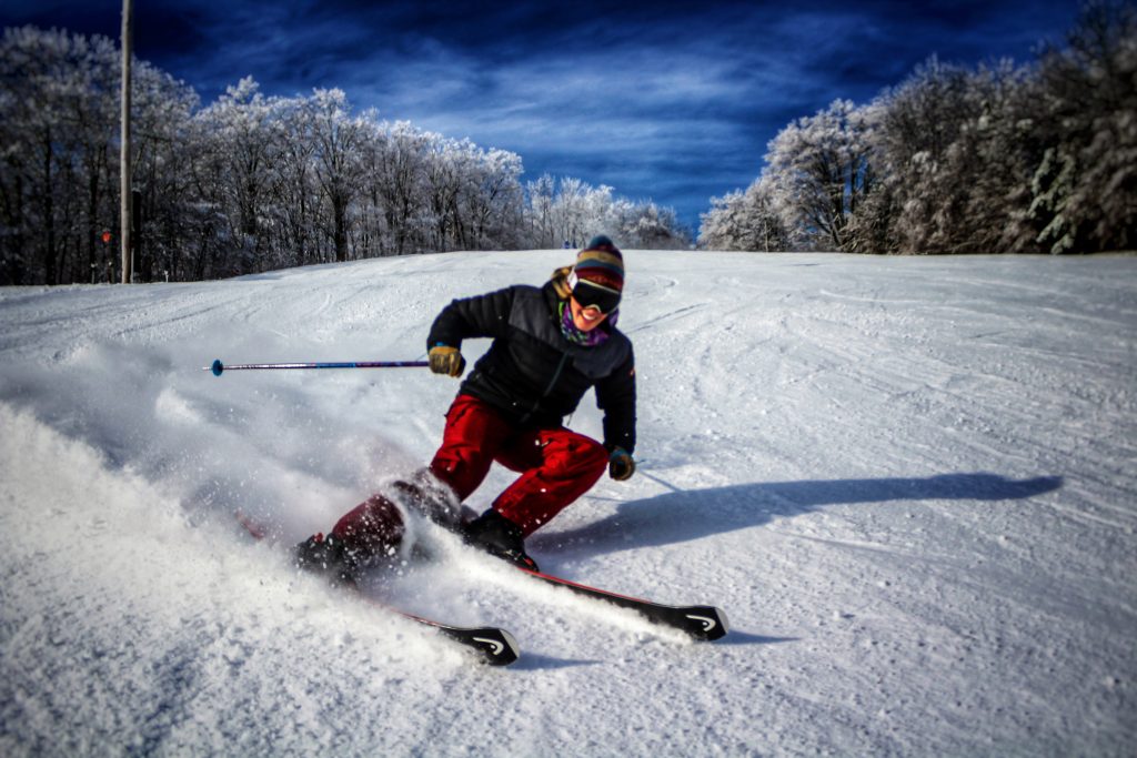 A skier gliding down a slope at Snow Ridge, amidst a lively winter sports scene in The Adirondacks, Turin, New York. The surrounding area is white with fresh snow, signifying a perfect day for skiing.