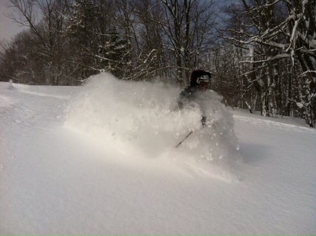 A snowmobiler rides past a chalet on the snowy slopes of Snow Ridge New York with skiers and snowboarders enjoying their winter sports in the background.