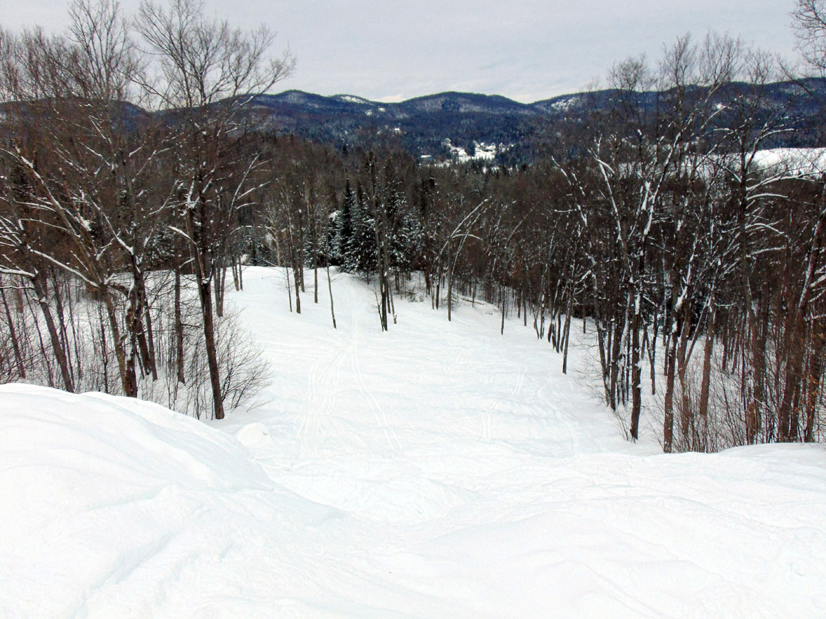 Winter scene at Mont Avalanche ski resort in Quebec, featuring a bustling ski scene with a ski lift transporting skiers uphill. A lone skier descends the slope, with a rustic chalet tucked away in the background.