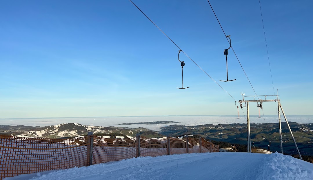A winter sports scene at Ebenalp-Horn, Eastern Switzerland, featuring a ski lift leading up the mountainside to a charming chalet. A surrounding ski resort creates a bustling winter sports centre.