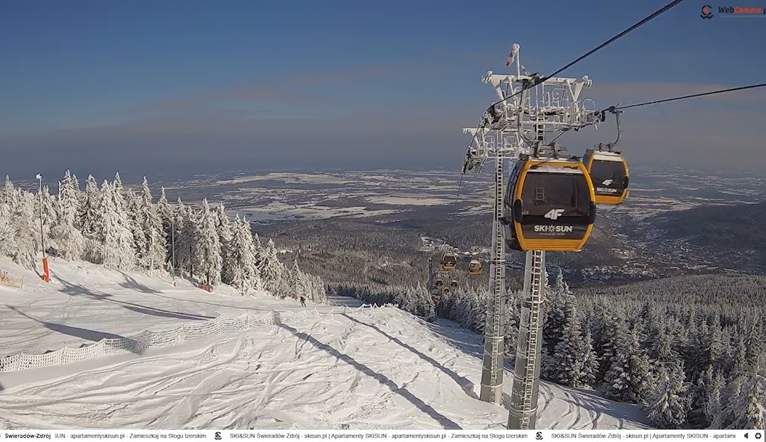 Scenic view of Świeradów Zdrój SKI & SUN Resort in Poland, featuring a ski lift traversing the snow-blanketed mountain, a skier gliding downhill, and a charming chalet nestled in the landscape.