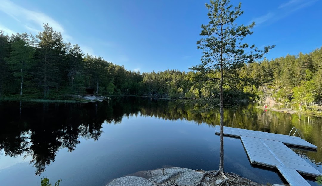 Sunlit view of a serene lake in Drammen, Norway, with a charming chalet and lodge nestled nearby. Gentle mountains frame the peaceful scene.