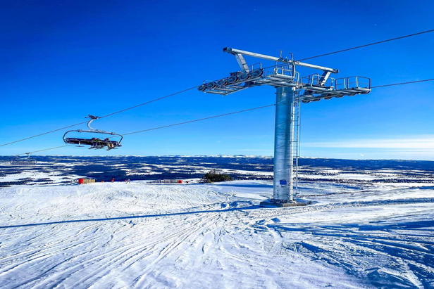 A picturesque view of Golsfjellet in Norway featuring a ski lift at a ski resort. Various skiers engage in winter sports amidst the serene snow-blanketed landscape. A small chalet is visible in the distance.
